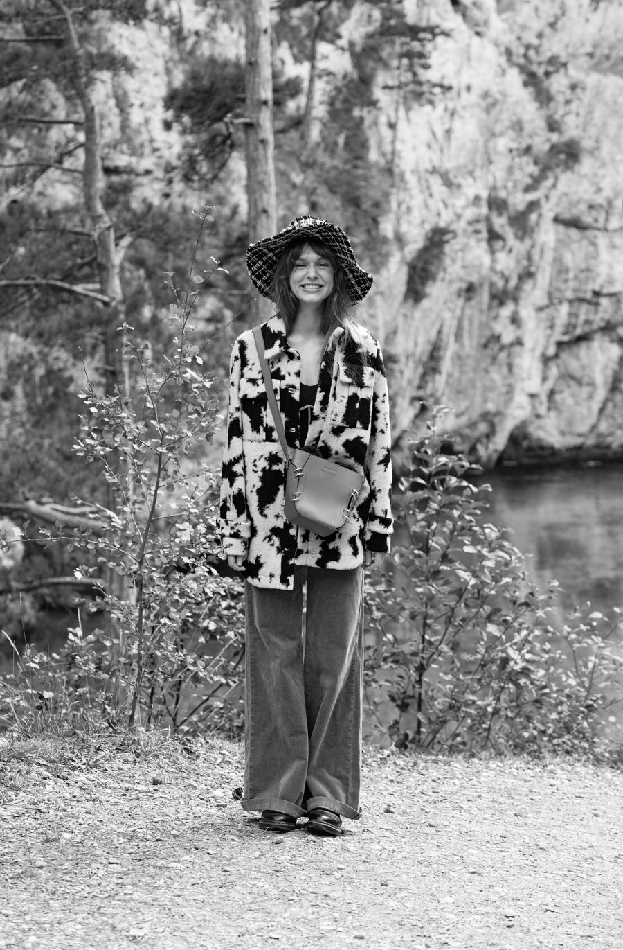 Black and white portrait of a woman in a patterned coat and hat, standing on a forest path.