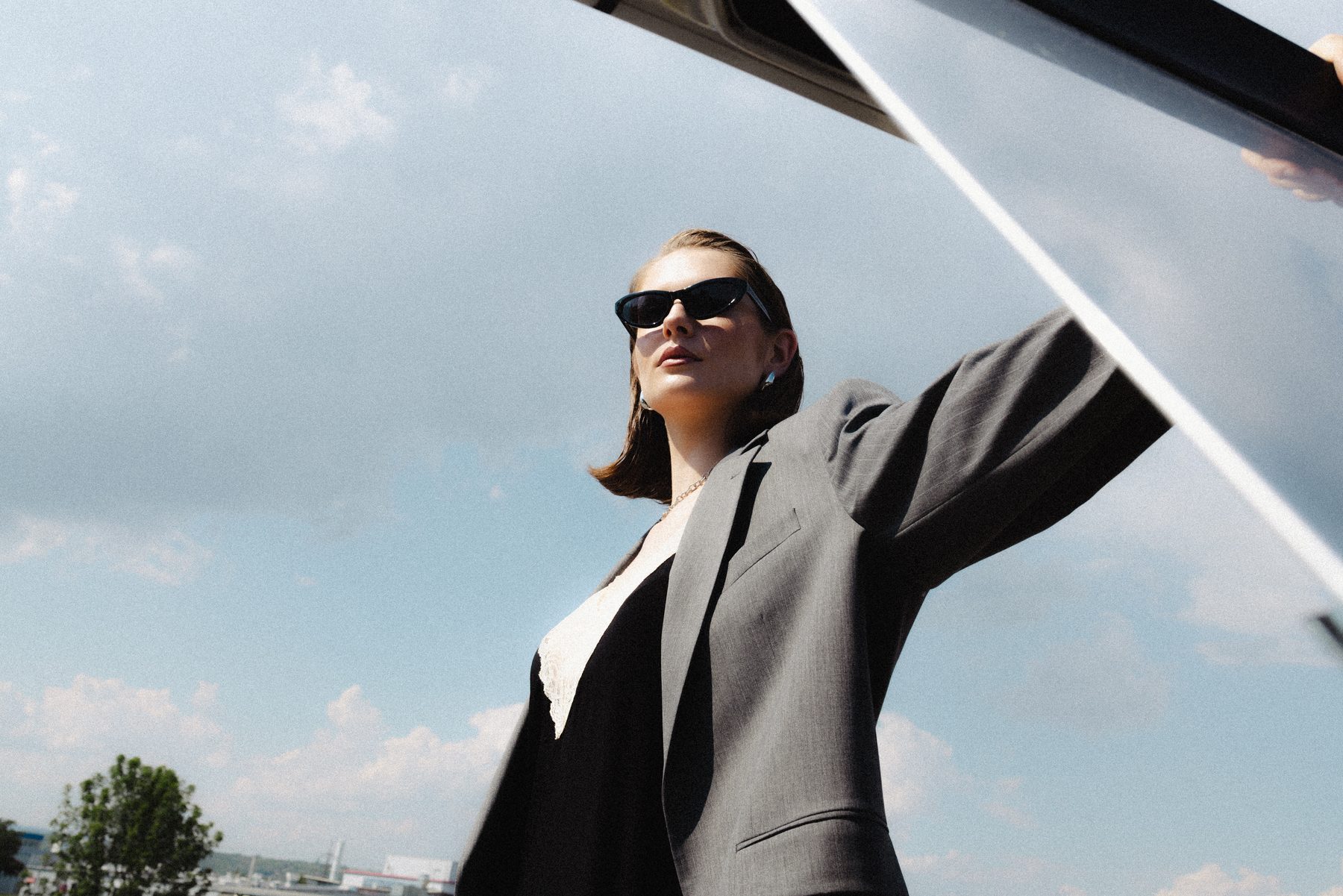 Portrait of a woman in sunglasses and a blazer, arm outstretched against a bright sky.