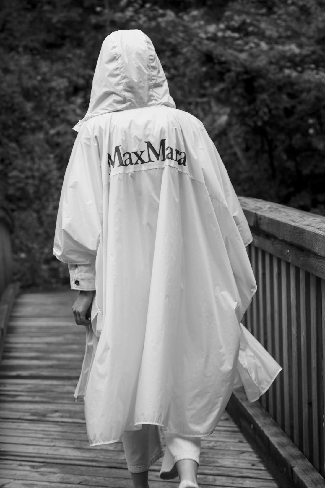 Person in a white MaxMara hooded rain cape walking along a wooden boardwalk, black and white version.